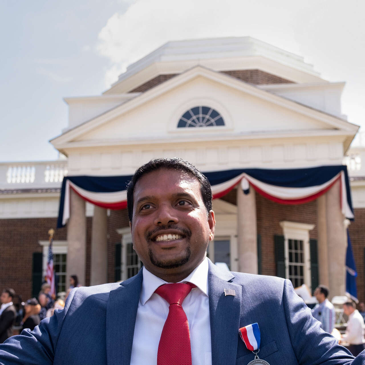 A man smiles while waving an American flag in each hand at a naturalization ceremony.